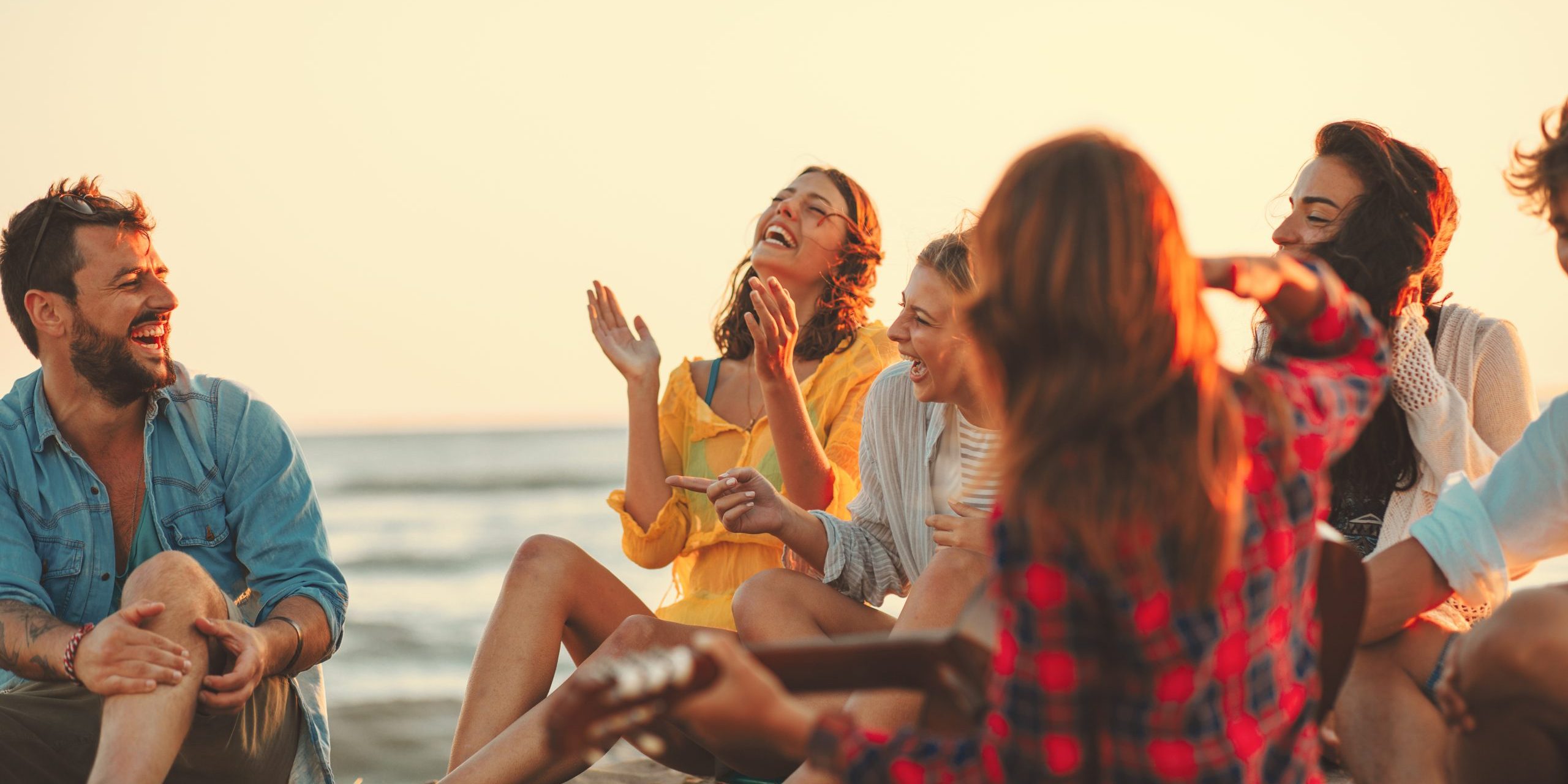 Happy friends sitting on the beach singing and playing guitar during the sunset Happy friends sitting on the beach singing and playing guitar during the sunset
